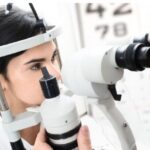 A imagem mostra uma mulher realizando um exame de vista em um consultório oftalmológico. Ela está sentada em frente a um equipamento usado para avaliar os olhos, chamado lâmpada de fenda. Um profissional da saúde, parcialmente visível, observa através do aparelho para examinar os olhos da paciente. Ao fundo, há uma tabela de letras usada para testes de visão. A cena transmite um ambiente clínico, limpo e bem iluminado.