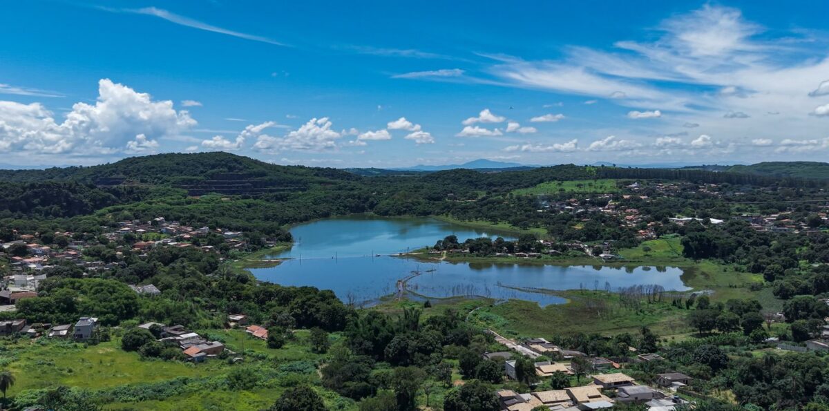 A imagem mostra uma vista aérea ampla do bairro Lagoa de Santo Antônio, em Pedro Leopoldo. O centro da fotografia é ocupado por uma grande lagoa de água azulada, cercada por áreas verdes e vegetação densa. Há pequenas ilhas naturais e áreas alagadas próximas às margens. Ao redor da lagoa, vê-se um conjunto de casas e ruas espalhadas entre árvores, formando o bairro. Mais ao fundo, morros verdes completam a paisagem, sob um céu azul vibrante com algumas nuvens brancas. A cena transmite tranquilidade e destaca a presença marcante da natureza no local.