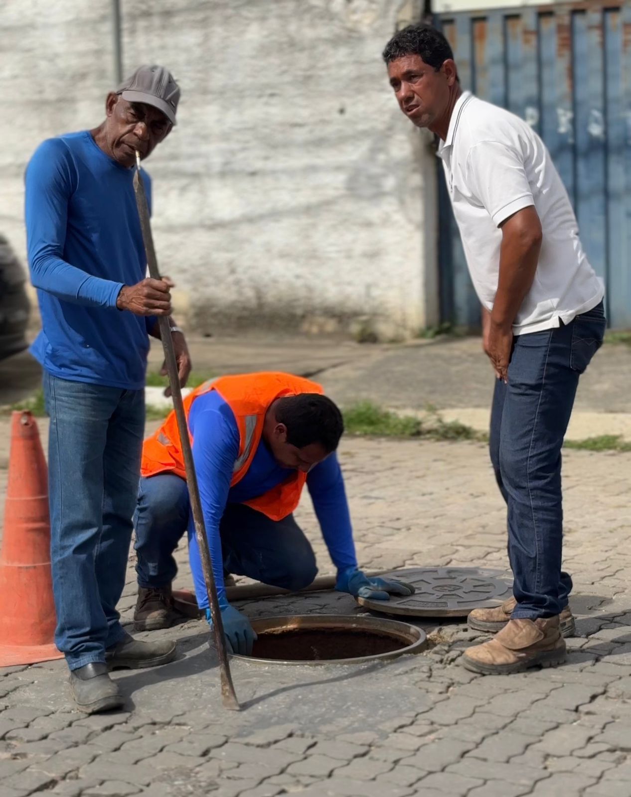 A imagem mostra três trabalhadores realizando serviço de manutenção em via pública. À esquerda, um deles segura uma barra de ferro usada para levantar a tampa de um bueiro; ele veste camisa azul, calça jeans, boné e botas. No centro, outro trabalhador está ajoelhado, usando luvas e colete laranja de segurança enquanto examina o interior do bueiro aberto. À direita, um terceiro homem observa o trabalho; ele veste camisa polo branca, calça jeans e botas claras. Ao fundo, há um muro cinza e um portão azul. Um cone de sinalização laranja está posicionado próximo ao local do serviço. A cena representa trabalho de manutenção urbana e cuidado com a infraestrutura da cidade.