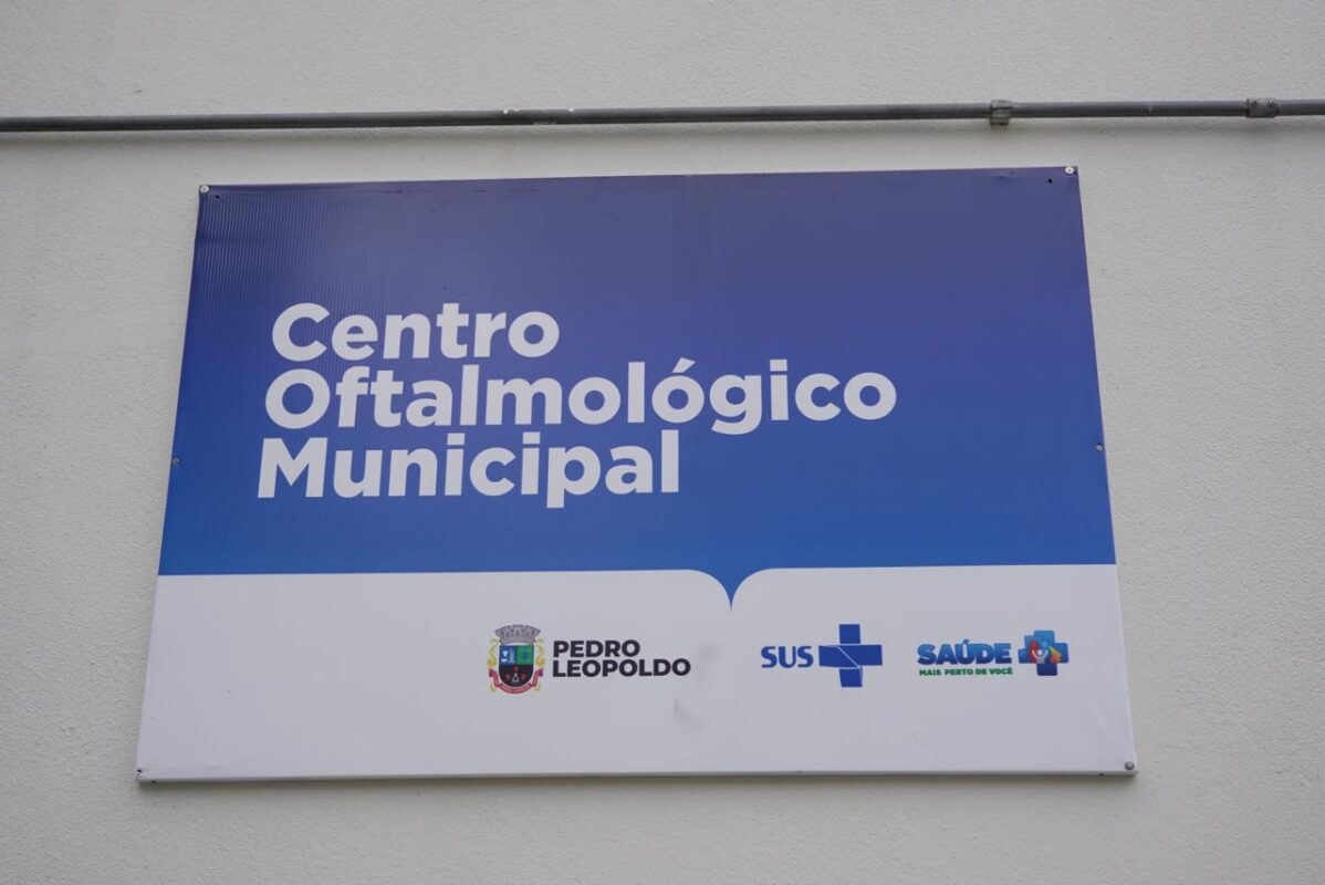 A foto mostra uma placa fixada na parede externa de um prédio. A placa tem fundo azul na parte superior e branco na parte inferior. No centro da área azul está escrito, em letras brancas e grandes: “Centro Oftalmológico Municipal”. Na área branca, na parte inferior, aparecem três logotipos: o brasão da Prefeitura de Pedro Leopoldo com o texto “Pedro Leopoldo”, o símbolo do SUS e o logotipo “Saúde – Mais perto de você”. A parede é clara e a placa está presa por parafusos nos quatro cantos.