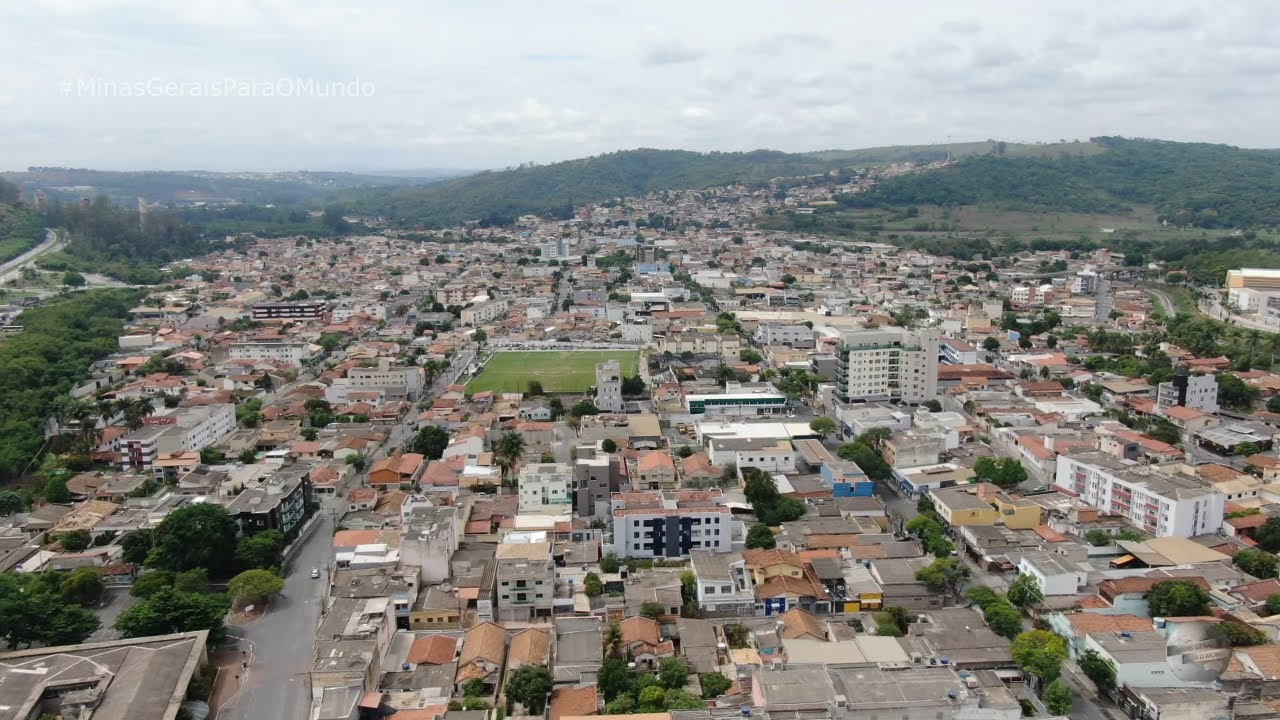 A imagem mostra uma vista aérea ampla da cidade de Pedro Leopoldo. Vemos um grande conjunto de casas, prédios e ruas formando o centro urbano, com construções de diferentes tamanhos e telhados predominantemente em tons de marrom e cinza. No meio da cidade aparece um campo de futebol verde, bem destacado entre as edificações. Ao fundo, há morros verdes e áreas de vegetação que cercam o município, dando sensação de horizonte aberto. O céu está parcialmente nublado, com luz suave. No canto superior esquerdo aparece a frase “#MinasGeraisParaOMundo”, escrita em branco e de forma discreta.